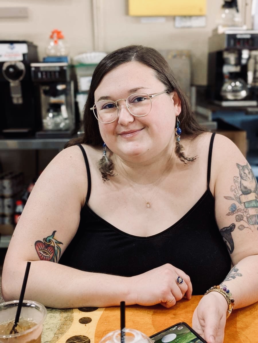 The artist, whom this introduction is about, is sitting and smiling softly at the camera. She is wearing a black top with spaghetti straps, feathered earrings, a ring, gold glasses, and colorful bracelets. She has brown hair swept behind her back and brown eyes. Some tattoos are visible as well as a drink in the bottom left corner. The artist appears to be in a cafe based on the background images of coffee pots and dispensers.