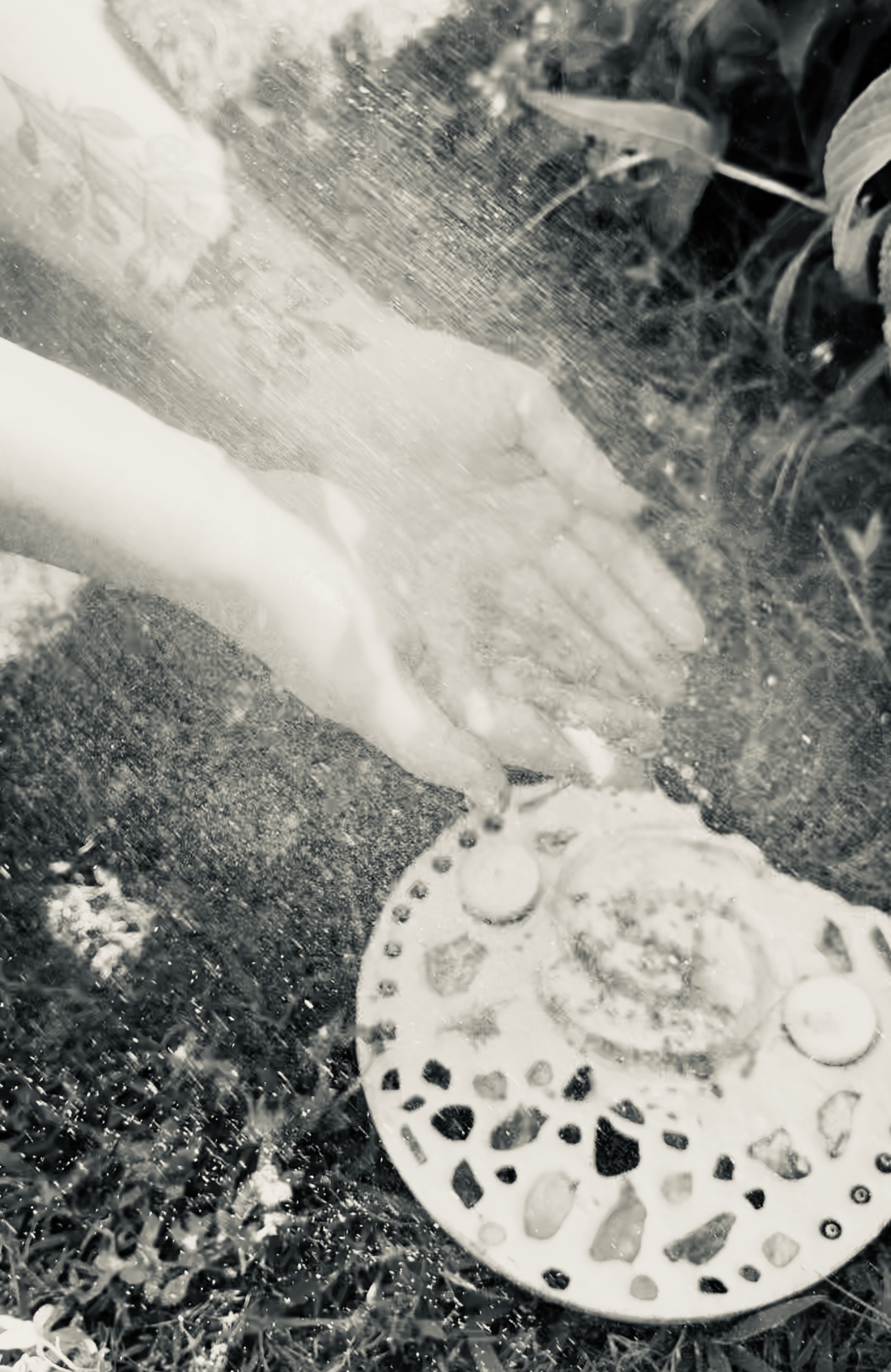 Black and white photo with arms coming from left side of photo. Hands are cupped together as water runs over them. The picture appears to be outside due to the grass and leaves in the background. There is a white circular object on the ground that is a stepping stone. It has rocks and candles embedded in it.